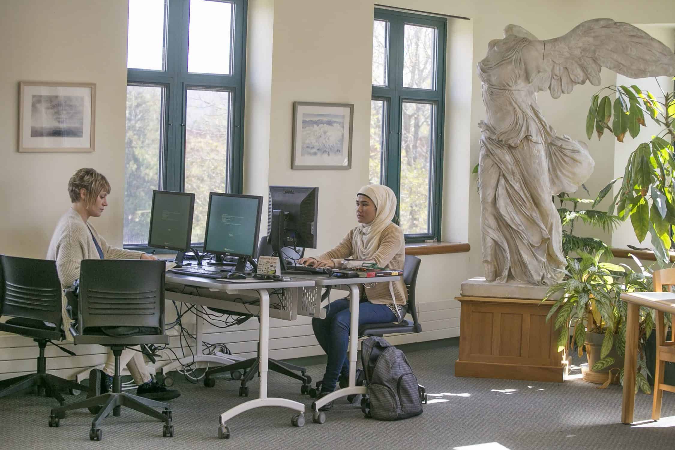 Students work at computers near a window with a statue in the background